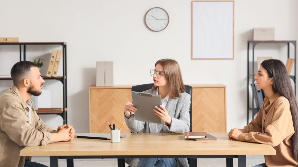 casal sentado cada um em um canto da mesa e mediadora no centro em audiência preliminar de conciliação de divórcio