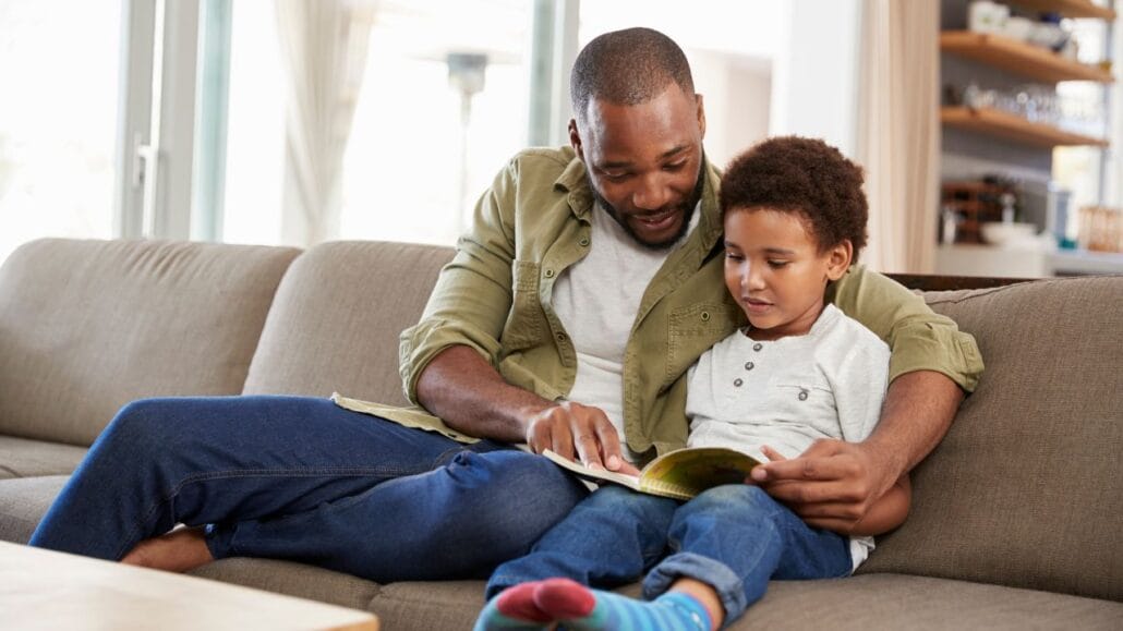 criança com livro aberto no colo. Ao lado pai ajudando a ler.