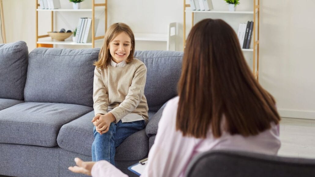 criança sentada no sofá e em primeiro plano psicóloga de costas a entrevistando para estudo psicossocial em ações com filho menor
