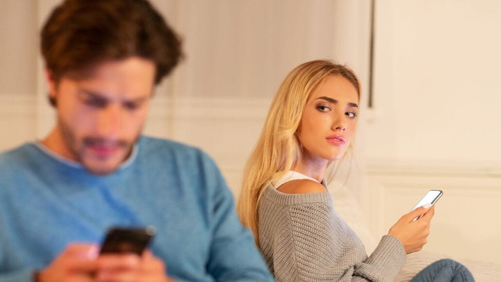 homem em primeiro plano com celular na mãe e mulher atrás também com celular na mão com expressão de desconfiada