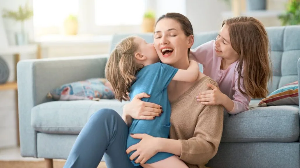 mulher sentada no chão em frente a um sofá abraçando uma criança e outra criança atrás com a mão em seu ombro
