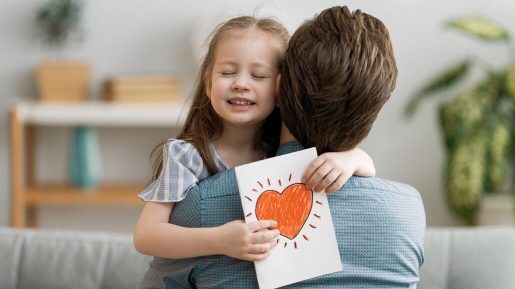 filha de olhos fechados abraçando pai com desenho de coração nas mãos 
