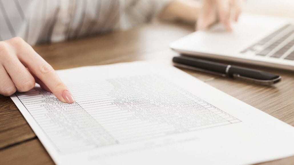 mão de mulher apontando para um documento de um lado e a outra mão mexendo em um notebook do lado de uma caneta analisando documentos